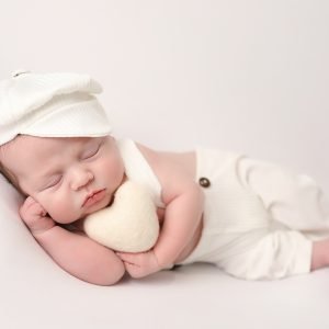 Newborn boy in dungarees and a cap holding a cream heart during a photoshoot