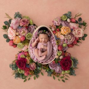 Newborn girl in a pink wrap surrounded by delicate flowers arranged in the shape of a butterfly.
