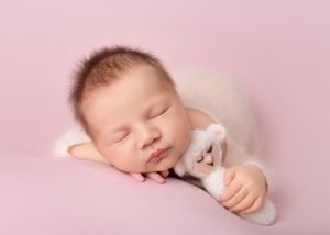 Newborn girl with a hairband on a baby pink background, holding a little lamb teddy bear at Daisy May Photography in County Armagh